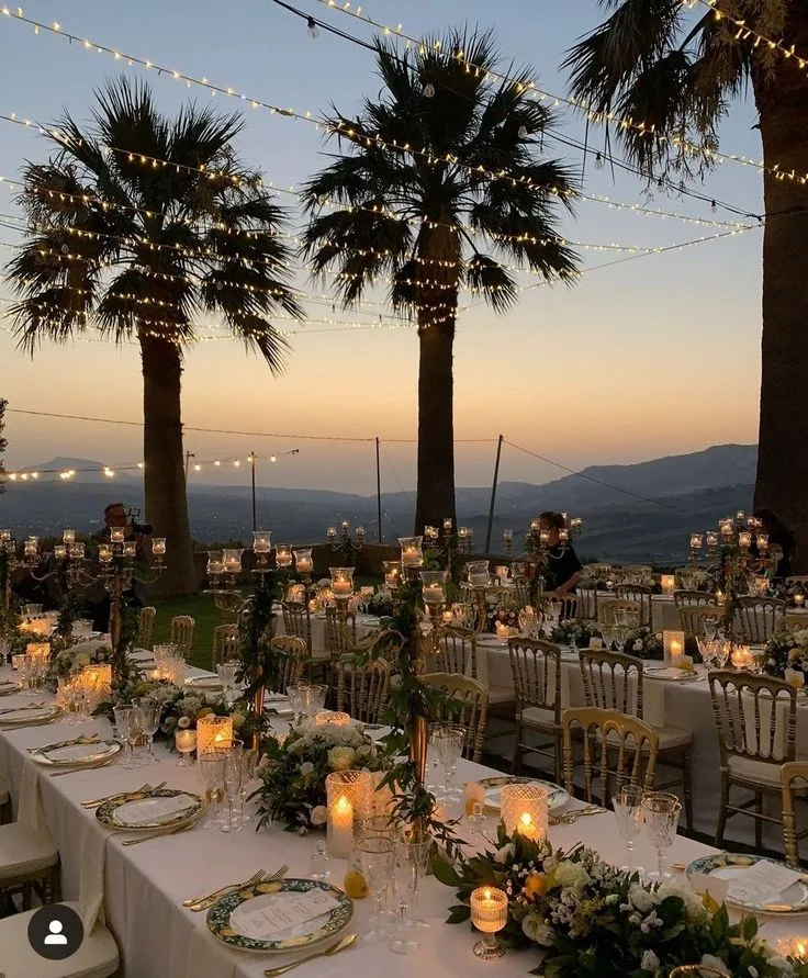 A long sit-down dinner table at dusk beneath string lights and palms